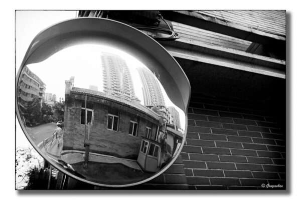 A large fish-eye mirror mounted on a corner of a brick building offers an interesting “rear-view” perspective on a street in Chengdu: in the foreground is a simple, one-story brick building with a tiled rooftop, while two enormous residential blocks tower in the background.