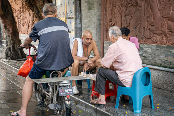 Three men sitting on low plastic stools play a game of Chinese chess on a sidewalk in Guangzhou, while another man, a passerby on a shopping-bag-laden moped, observes their game.