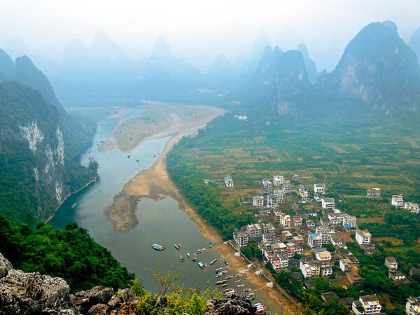 A bird’s eye view of heavily forested, dramatic karst peaks along the Li River. Various colorful water craft ply their way along the river, and on the right bank, there is a small cluster of modern, three- and four-story residential buildings.