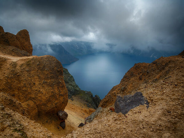 Viewed from atop a peak of black and brown volcanic rock, an enormous lake, formed from a volcanic crater, stretches into the distance, with low dark clouds shadowing its surface.