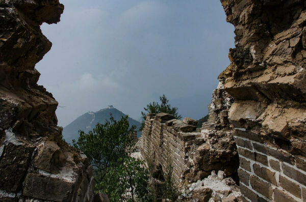 A view from inside a crumbling portion of the Great Wall reveals another section of the wall snaking up a distant hill, atop which sits a guardhouse, and beyond that, misty clouds.