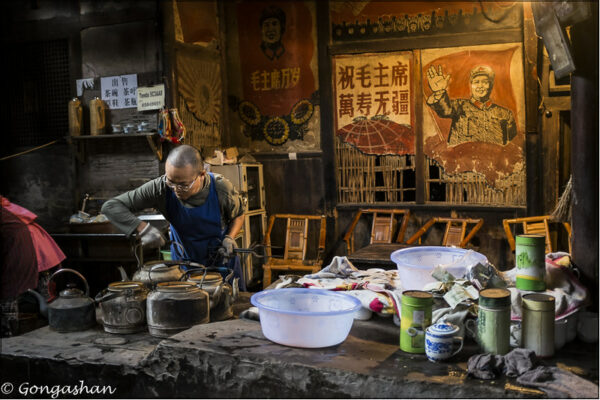 A man with a shaved head, glasses, and a blue apron peers at several metal teapots on a stove at a teahouse in Chengdu, Sichuan Province. The workspace in front of him is littered with plastic basins, ceramic cups, and metal tea tins, while the wall behind him is decorated with a number of Cultural Revolution-era propaganda posters with revolutionary slogans and images of Mao Zedong.