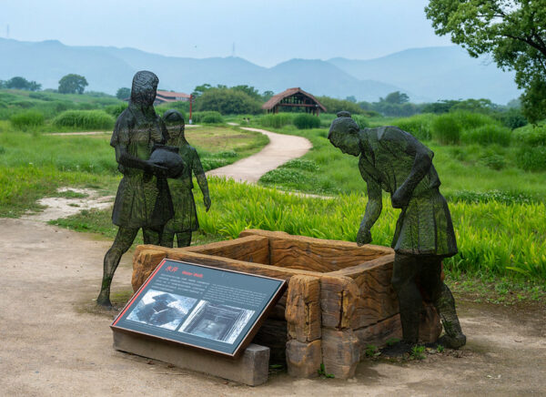 At the Archaeological Ruins of Liangzhu City, a UNESCO World Heritage Site near Hangzhou, three semi-transparent black metal sculptures of a Neolithic man, woman, and child cluster around a rectangular wooden well. Behind them, a dirt path winds through a meadow toward a simple structure with thick wooden beams and a thatched roof.
