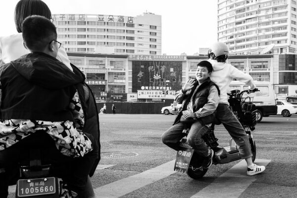 At a large intersection in Qingdao, an adolescent boy riding on the back of a scooter smiles and waves at a friend, a boy of about the same age, who is riding on the back of another scooter.