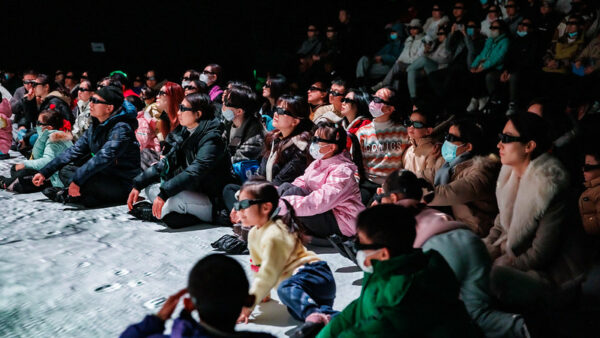 The bleachers and floor are filled with museumgoers of all ages, raptly observing an exhibition and wearing what appear to be VR goggles. Nearly everyone is bundled in coats and sweaters, and some are wearing surgical masks.