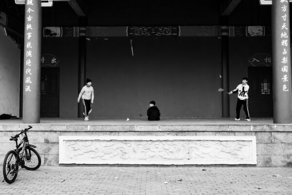 Two boys dressed in athletic clothes play a game of badminton on a raised platform between the pillars of a traditional Chinese building adorned with calligraphy. Another boy, dressed in a dark shirt, sits on the ground between the two boys who are playing.