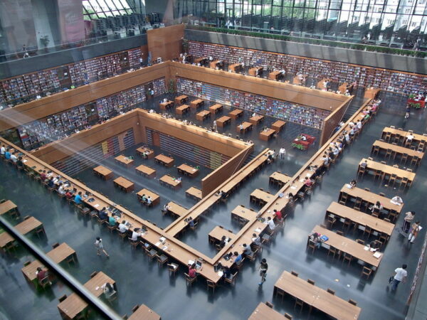 A bird's eye view of the interior of the National Library of China reveals an open atrium, multiple floors lined with bookshelves, and many patrons sitting at wooden reading tables.