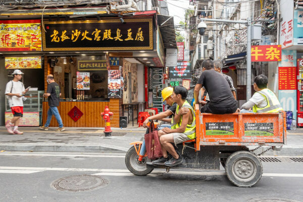 Five construction workers are crowded onto the seat and back-bed of a small, orange, three-wheeled motorized cart traveling down the street of what appears to be a busy commercial district in Guangzhou.