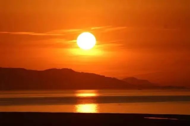 Photo of a beautiful sunset shows the sun as a white orb against a background of orange and red clouds, setting over some hills and a body of water.