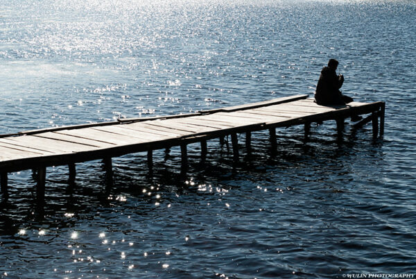 A man sits alone at the end of a narrow wooden dock that extends into dark blue waters. Sunlight glints off tiny ripples on the surface of the water.