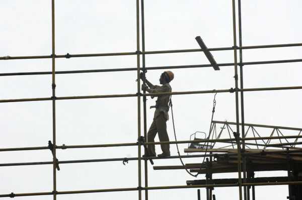 Against an overcast sky, a worker wearing tan clothing and an orange hard hat stands on a narrow beam of some high scaffolding. He is connected to the scaffolding only by a thin rope attached to a harness at his waist.