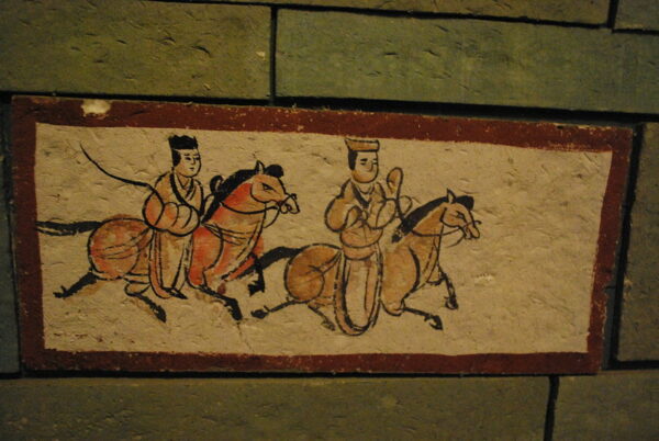 A Han Dynasty cave painting on a rectangular brick uses bold black calligraphic strokes and shades of tan, yellow, orange, and red to depict two horses and their riders, who are attired in headdresses and long robes.