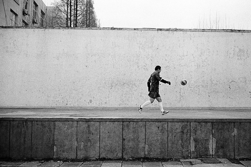 A man dressed in soccer gear kicks a ball against a white wall atop a raised platform. Behind the wall, a concrete building, a few trees, and an overcast sky are visible