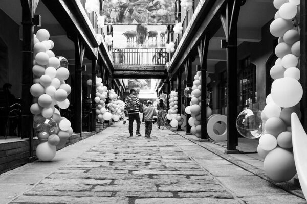 A father and son, and a few other passersby, walk through a breezeway lined with wooden pillars decorated with enormous bunches of festive balloons, some opaque white and some clear.