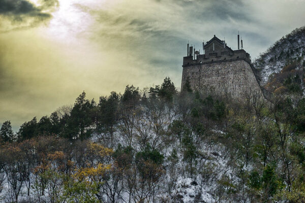 A stormy sky looms over a lightly forested hillside dusted with snow. Atop the hill, a gray brick former guard tower of the Great Wall of China bristles with cell phone antennae.