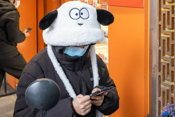 A woman looks down at her cell phone as she stands outside a bright orange doorway on Haizhu S Road in Guangzhou. She is dressed in a black puffa jacket, blue surgical mask, and fuzzy black-and-white hat shaped like a dog with floppy ears.