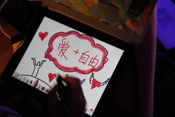 An illustration on a digital pad device shows the outline of a person with their hands in the air, hearts, hearts with wings, and in a cloud at the center, the message “Love and Freedom.”