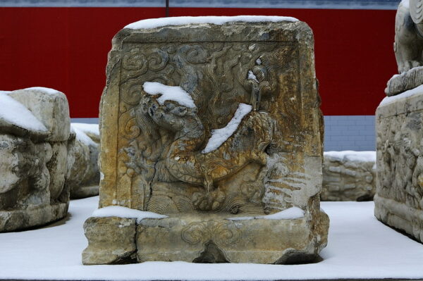 Against a bright red wall and white snowy ground, snow has settled into the crevices of a stone ornament carved with a fanciful creature, making it appear that the creature is wearing a snowy white hat and cloak.