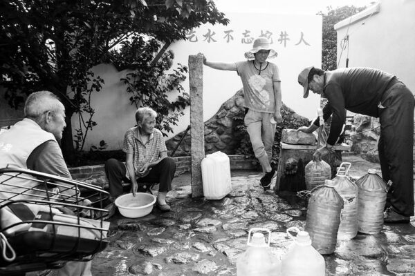 A group of four people are gathered around a well. At right are a middle-aged man and woman filling large plastic bottles with water. At left are an older man with a bike or motorbike, and an older woman with a plastic basin full of water.