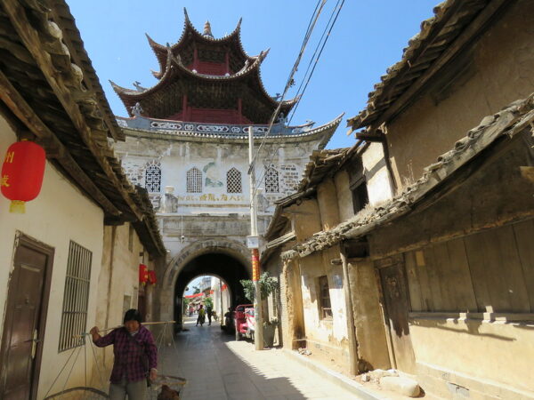 A narrow alleyway is dominated by a tall, elaborate city gate with a rounded entrance and towering, red, pagoda-like structures atop it. There are a small number of passersby in the alleyway, including a woman balancing two baskets from a bamboo pole she carries over her shoulder.