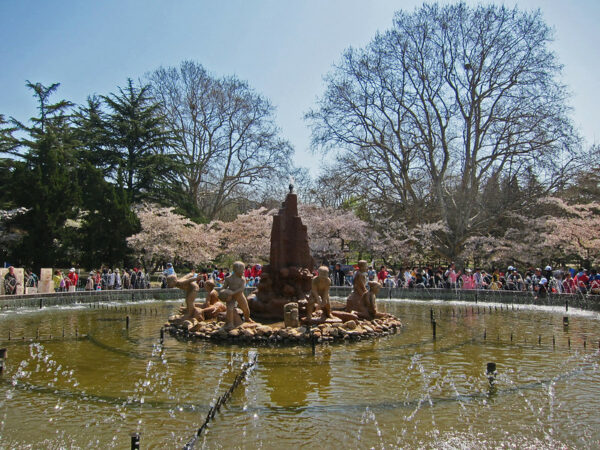 Against a backdrop of tall trees and early spring foliage, a group of brightly dressed people surround the margins of a large circular fountain with a sculpture in the center depicting a group of children playing.