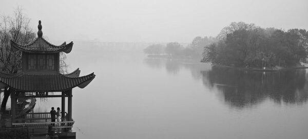 A misty image of Nianjia Lake (Chansha, Hunan province) in the rain. On the left shore is a traditional Chinese pavilion with two people standing inside it; on the right shore are outcroppings of trees, blurred in the distance.