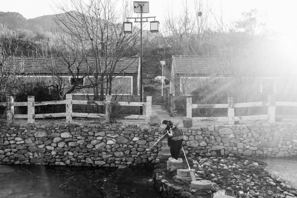 A woman stands on a cobblestone and stone-wall-reinforced riverbank, cleaning or wringing out a mop over the river. Behind her are some wooden fences, trees, and two single-story brick houses.