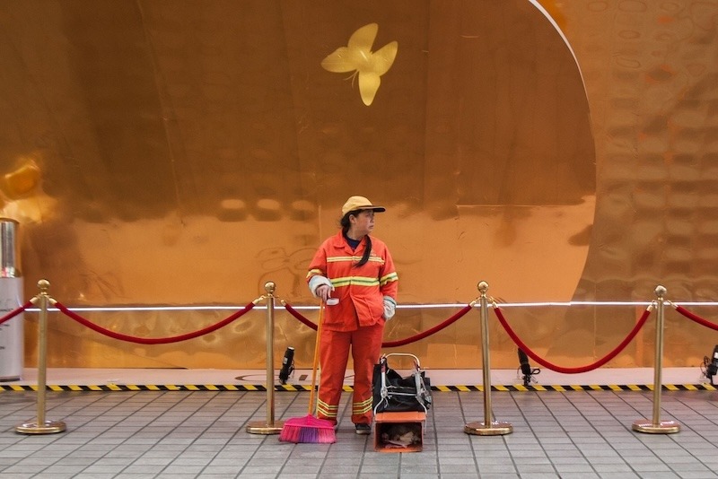 A female sanitation worker dressed in a bright orange and yellow uniform with a cap stands in front of a golden wall protected with a red-velvet guard-rail