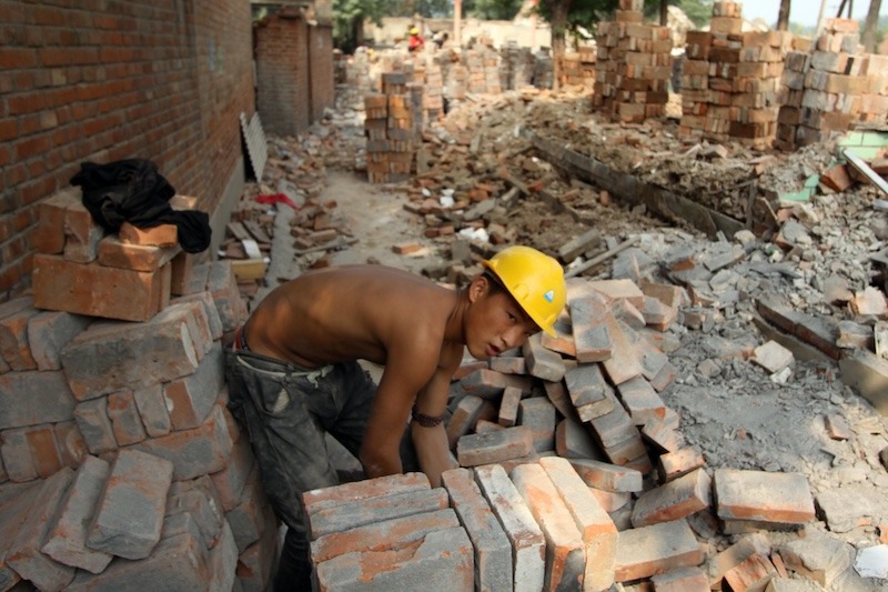 A bare-chested young man wearing army-green trousers and a bright yellow hard hat bends over a pile of dusty red bricks.