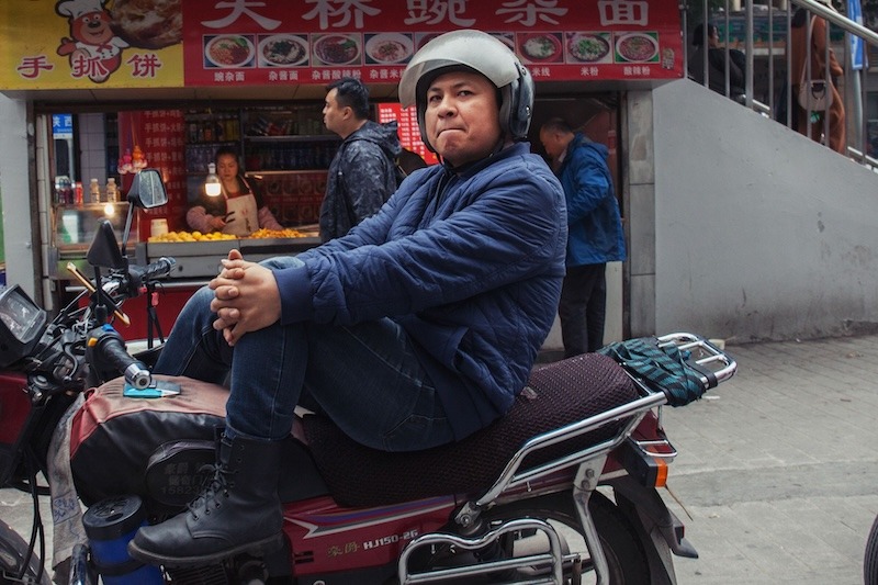 Parked in front of a small noodle stall, a driver wearing a helmet, padded blue jacket, blue jeans, and black leather boots leans back in the seat of his motorcycle and and relaxes.