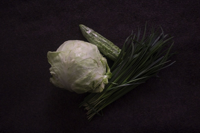 A head of cabbage, a bunch of chives, and a bitter melon against a black backdrop.