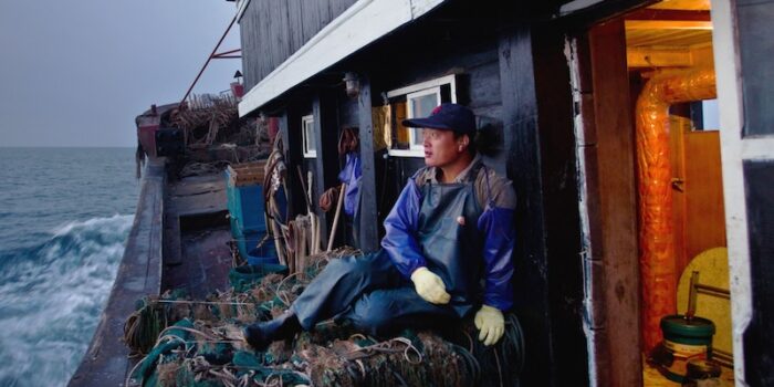 A young fisherman dressed in warm clothes and a rubber apron, sleeve protectors, and gloves, sits on a pile of fishing nets on the side of a trawler out at sea.