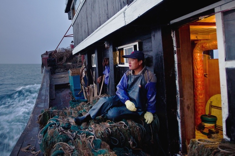 A young fisherman dressed in warm clothes and a rubber apron, sleeve protectors, and gloves, sits on a pile of fishing nets on the side of a trawler out at sea.