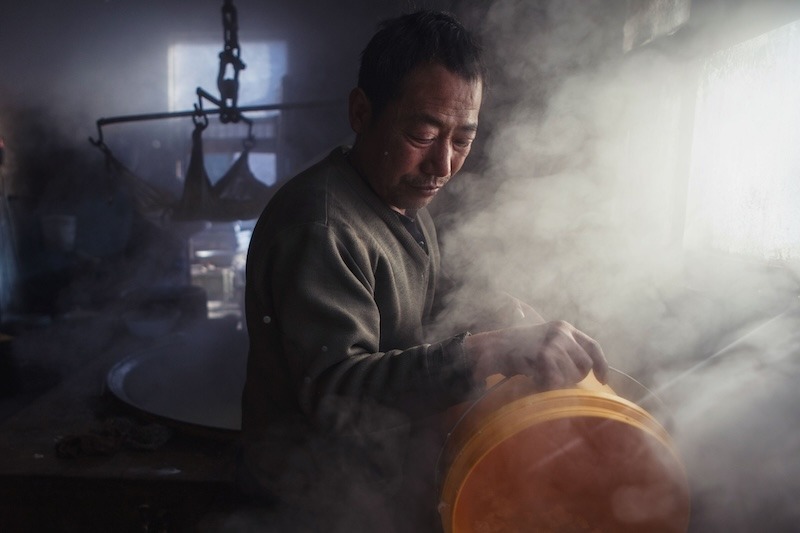 Surrounded by steam, a man in a khaki sweatshirt pours something from a bright yellow plastic bucket.