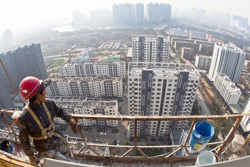 A slim young man in khaki clothes and a red hard hat looks down from a very high scaffolding: from his vantage point, the ten- and -twenty story buildings below him look quite small.