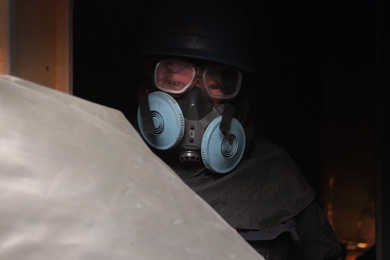 A worker wears a black hard hat, clear goggles, and a full respirator.