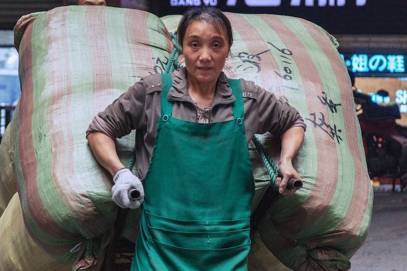 A woman wearing a bright green apron and white cotton working gloves pulls an enormous load of something encased in woven bags on a metal trolley behind her.