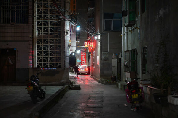 A narrow alleyway with residential buildings on both sides is lit by a red neon sign for a small guesthouse and the red tail lights of a car stopped at the end of the alley. In the glow of the tail lights, a man holding a ladder is visible, as are two parked mopeds in the foreground. 