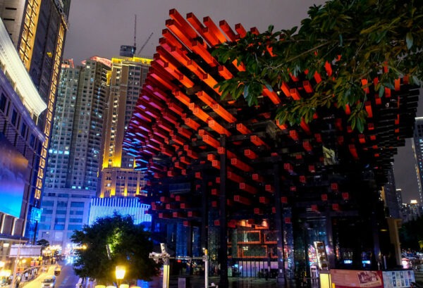 Against an evening cityscape of towering skyscrapers and brightly lit buildings, a complex lattice of red beams forms the entrance to a theater featuring Peking Opera in the city of Chongqing, Sichuan Province.