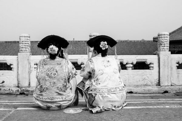 A photo, taken from behind, of two women—one of whom is looking at her cell phone—sitting side by side in what appears to be Beijing’s Forbidden City. The women are dressed in silk brocade gowns with elaborate traditional headdresses and fans, much like the empresses and imperial concubines of old.