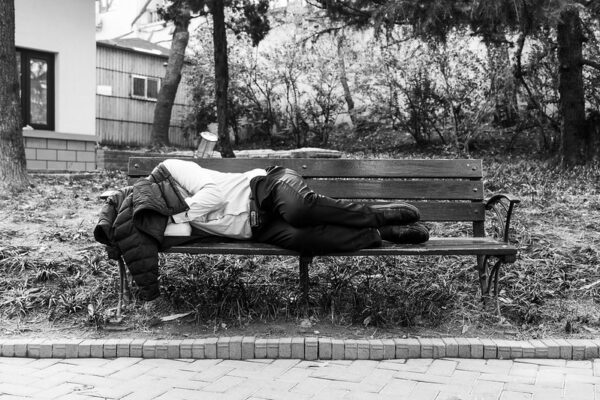 A man wearing black slacks, a white button-down shirt, and dress shoes with a dark-colored down jacket draped over his head, takes a nap on a park bench amidst the trees.