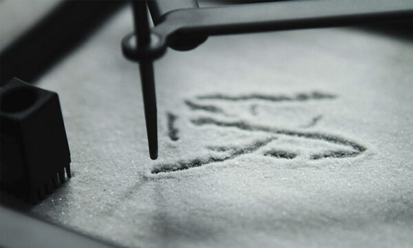 Close-up view of a mechanical apparatus with an attached metal stylus inscribing a Chinese character in a thin layer of white sand.