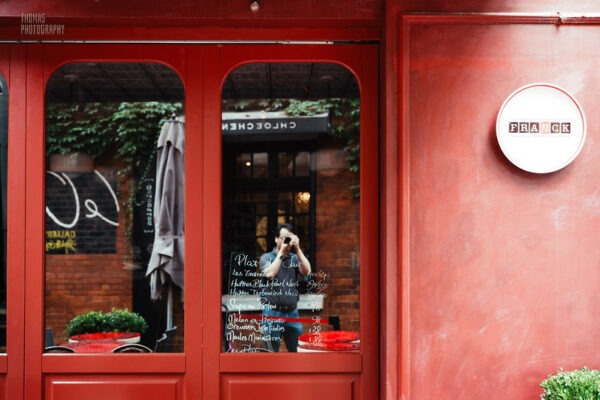 A man pausing in the street to take a photograph is partially visible in a reflective window of a charming building with red walls and red wood-framed windows.