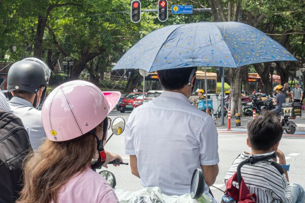 A group of pedestrians and cyclists wait for the light to turn green at a busy intersection in Guangzhou. Among the waiting crowd is a young boy with a striped shirt and red backpack, a woman in a pink shirt and pink helmet, a tall pedestrian in a white shirt with a blue sun umbrella, and an older man with a black helmet and visor.