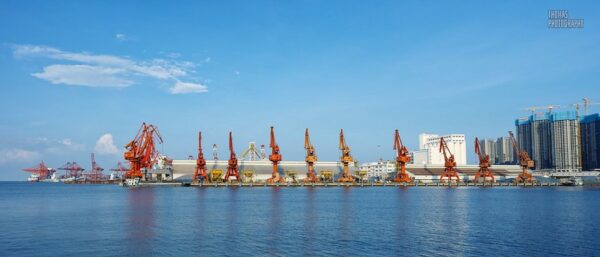 Shenzhen’s Shekou Port is dotted with over a dozen enormous metal cranes in shades of red, orange, and yellow. The waters surrounding it are a brilliant blue, and the sky above a slightly lighter shade of blue, with a few wispy clouds floating overhead.