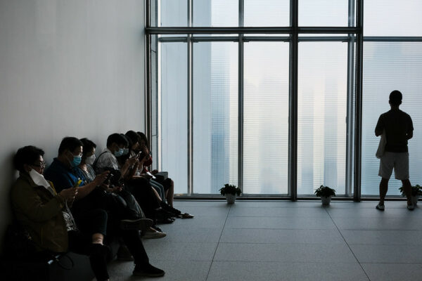 A man stands in silhouette before a bank of floor-to-ceiling semi-opaque windows that reveal a vague cityscape of skyscrapers outside. To his left, about a half-dozen people—some masked, some not, and many looking at cell phones—sit in a row of chairs against the wall, waiting for something.
