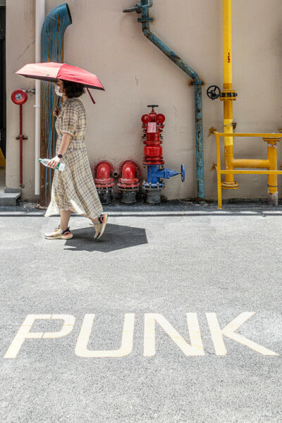 A woman wearing a plaid dress and tennis shoes and carrying a red sun umbrella walks past a building whose various exterior pipes and valves are painted in eye-catching shades of yellow, red, and blue. White painted English text on the asphalt reads, “PUNK.”