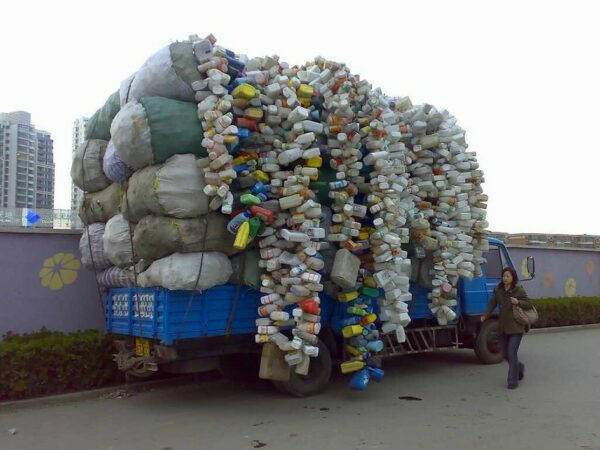 A large blue flatbed truck is intricately, almost sculpturally, loaded with enormous bags and very long strings of recyclable plastic bottles and containers that tower to a height at least three times as high as the truck’s cab. A pedestrian passes by the truck, parked next to a wall with a colorful lavender mural, without a curious glance.