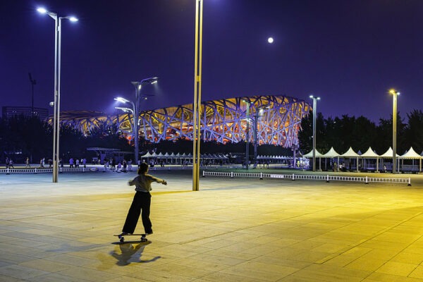 A skateboarder in a white t-shirt and loose black trousers practices some moves beneath a full moon and the tall, bright lights of a vast and mostly empty public square. Beijing’s brilliantly lit “Bird’s Nest” stadium is visible in the background.