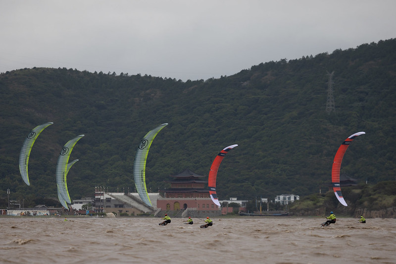 About a half-dozen formula kite racers dressed in bright yellow skid across the surface of the ocean, their crescent-shaped kites in shades of red and blue, gray and yellow visible in the air above them. On the shore behind them is a dark red three-storied traditional Chinese building, and a deep green forested hillside.